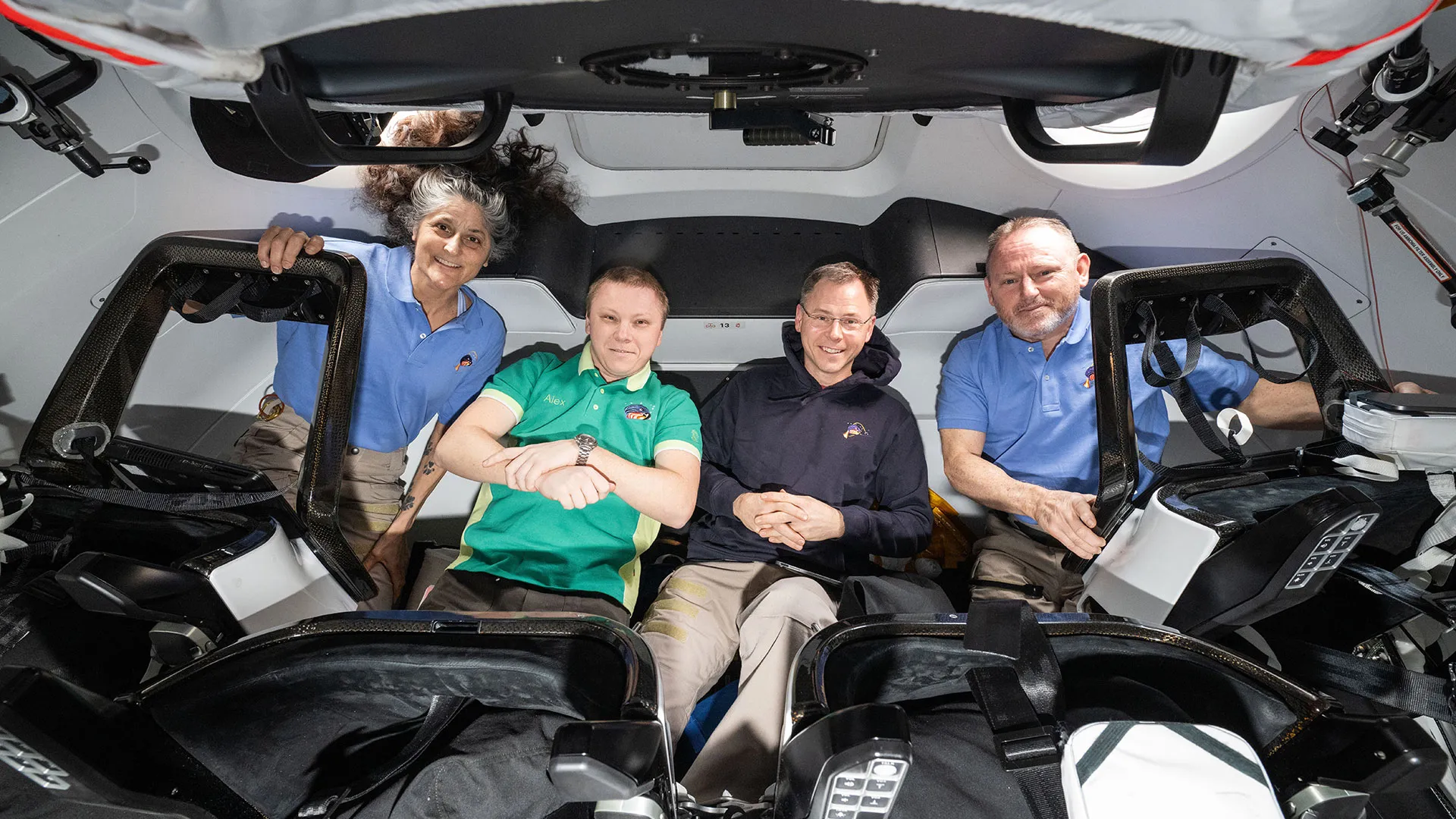 Drei Männer und eine Frau in Polo-Shirts und einer Kapuzenjacke schweben in einem Raumschiff für ein gemeinsames Foto.