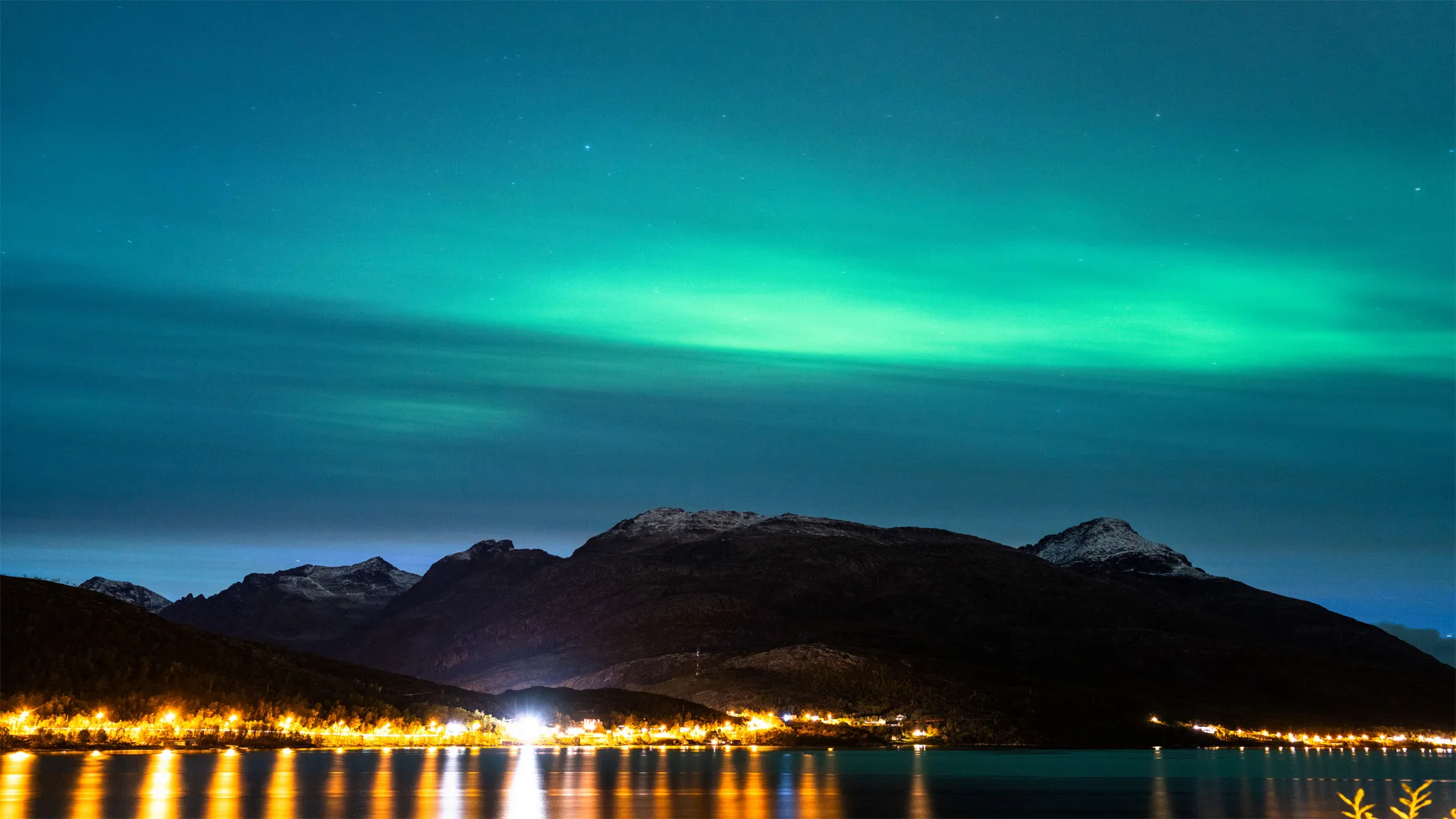 Das Nordlicht über einer Bergkette mit hellen Straßenlaternen im Vordergrund.
