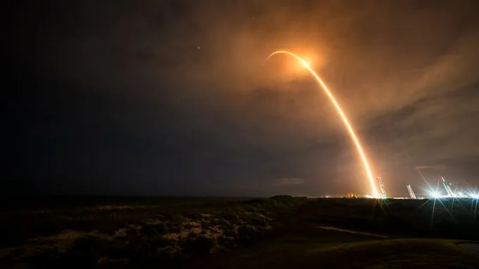 Ein Raketenstart zeichnet einen orangefarbenen Bogen in den dunklen Nachthimmel auf diesem Langzeitbelichtungsfoto.